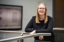 Landscape colour photograph of Marge Ainsley. Marge has long blonde hair, is wearing glasses and a black top. She leans on a railing and smiles.