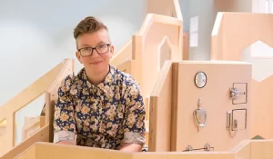 Photograph of Margaret Middleton, freelancer in LGBTQ inclusivity. Margaret has short hair styled into a quiff, wears glasses and a patterned shirt. They are sitting in front of an indoor museum play area with a playhouse.