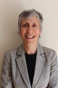 Photograph of woman from head to chest, looking at camera. Caroline has short grey hair and is smiling. She wears a grey blazer with black top underneath.