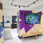 A photograph of an exhibition space with white walls and ceiling. In the centre stands a tall purple and brown box with a display screen inside. the screen is visible to those who stand in front of it. More interpretative panels and another screen can be seen on the wall on the background